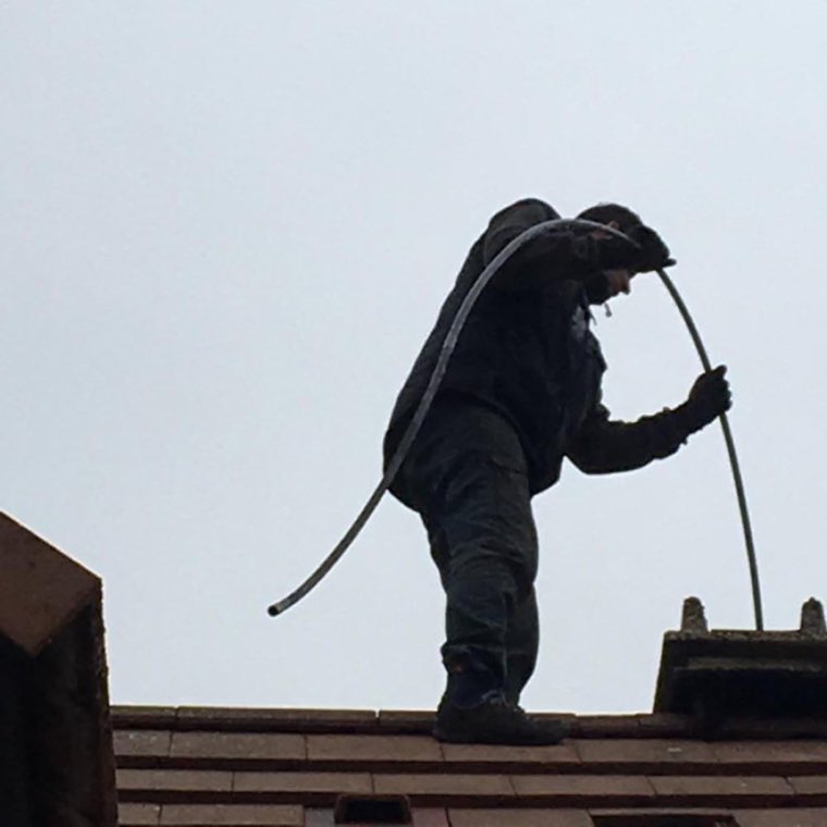 Ramoneur en tenue de travail sur le faîtage d'un toit, canne de ramonage à la main